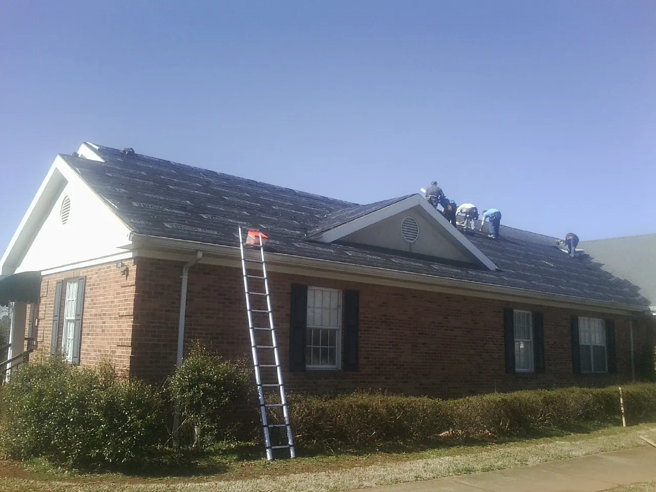 Crew tearing shingles off a brick ranch roof with a ladder set — Emergency Roof Repair work by our crew in Clear Brook