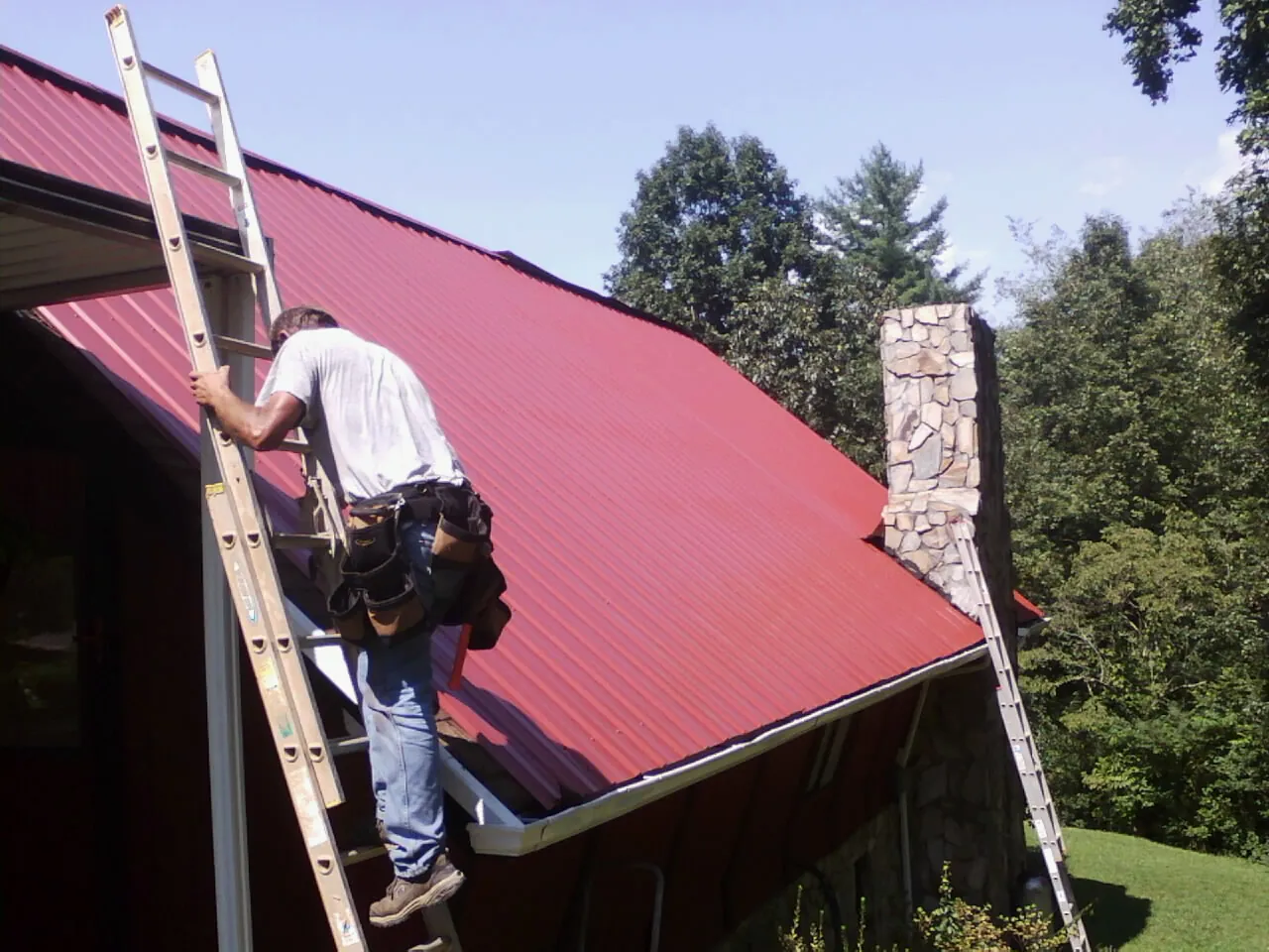 Roofer on a ladder beside a red metal roof and stone chimney — Soffit & Fascia Repair by Roofing Brothers in Clear Brook